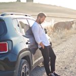A man in a white shirt leans against a car on a sunny rural dirt road. Simplicity and solitude.